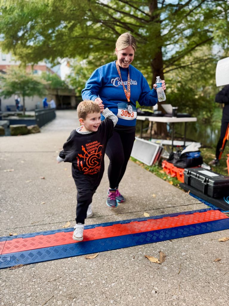 a smiling young boy in a gahanna community chorus spooky shuffle shirt crossing the finish line hand in hand with a runner wearing her medal